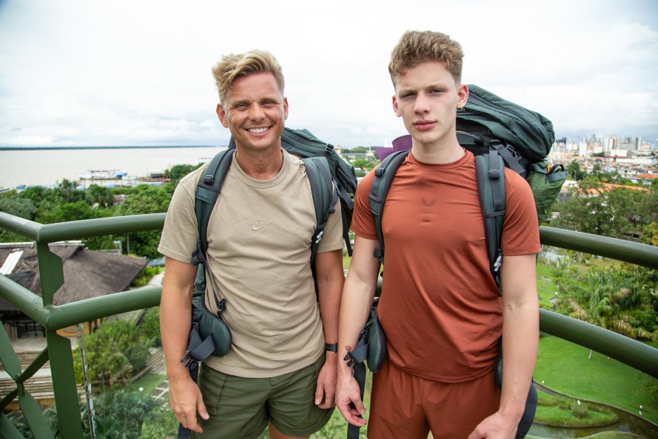 Jeff Brazier and his son Freddy, wearing backpacks, on a balcony overlooking a river and city.