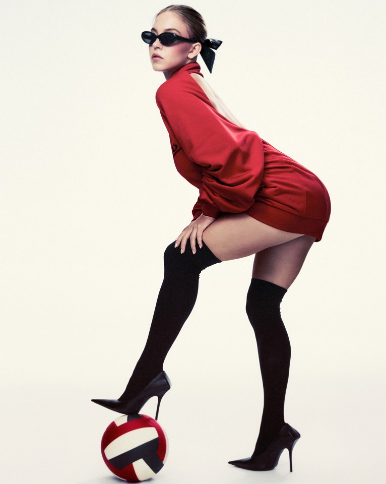 Sydney Sweeney in a red dress and black heels, balancing on a volleyball.
