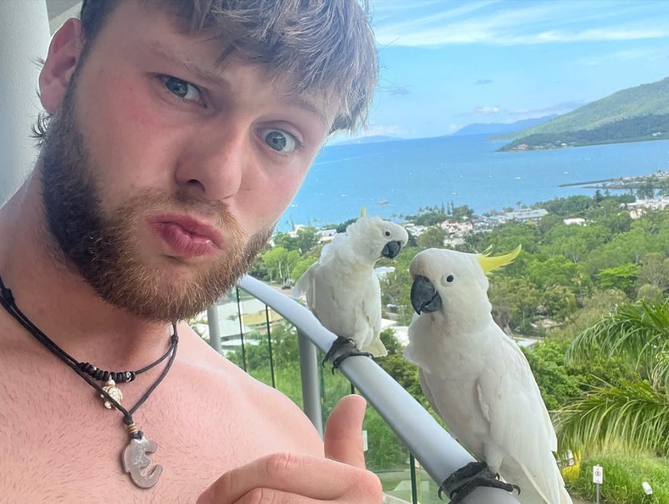 Man with two white cockatoos on a balcony overlooking the ocean.