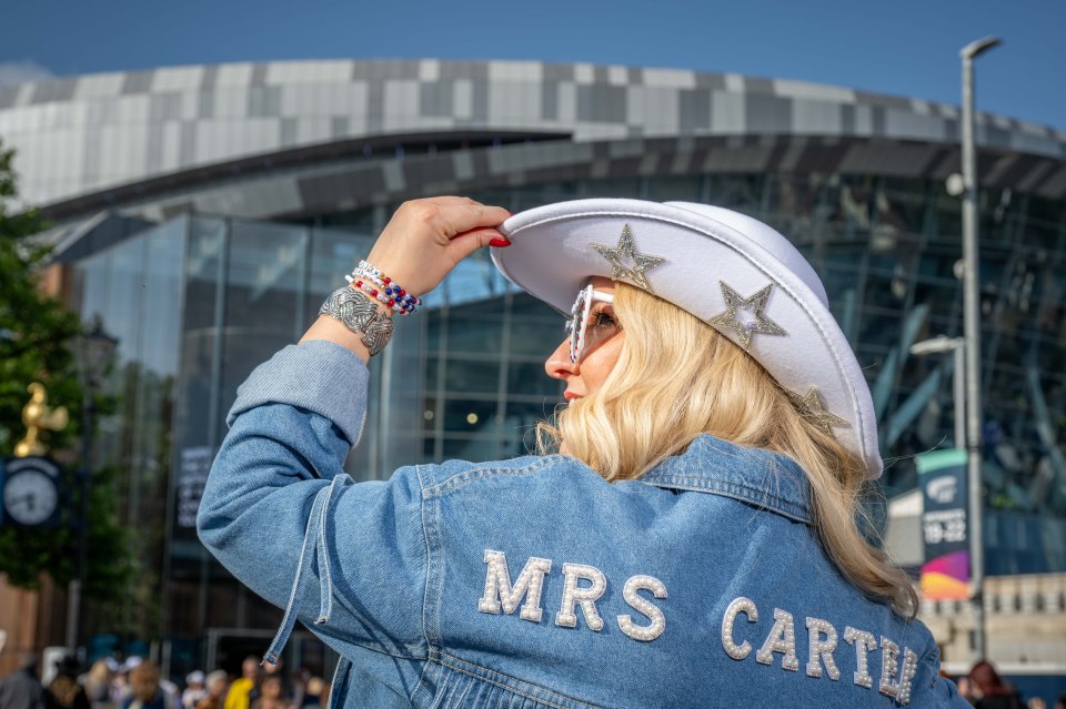 Woman in a denim jacket with "Mrs Carter" on the back, wearing a white cowboy hat, adjusting her hat outside a stadium.