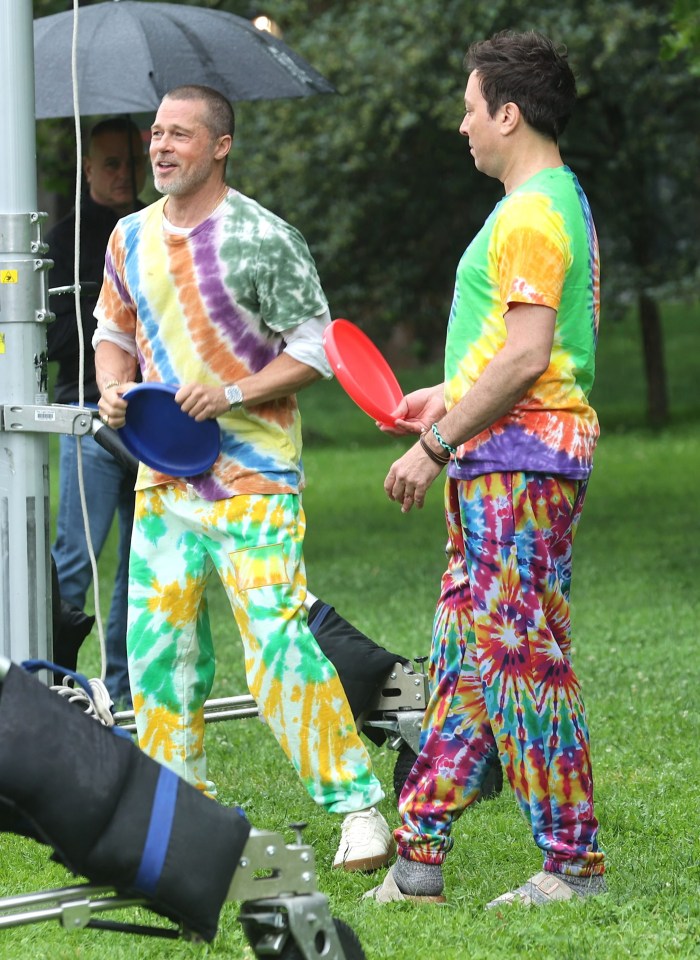 Brad Pitt and Jimmy Fallon filming a skit in a park, wearing tie-dye shirts and pants, playing frisbee.