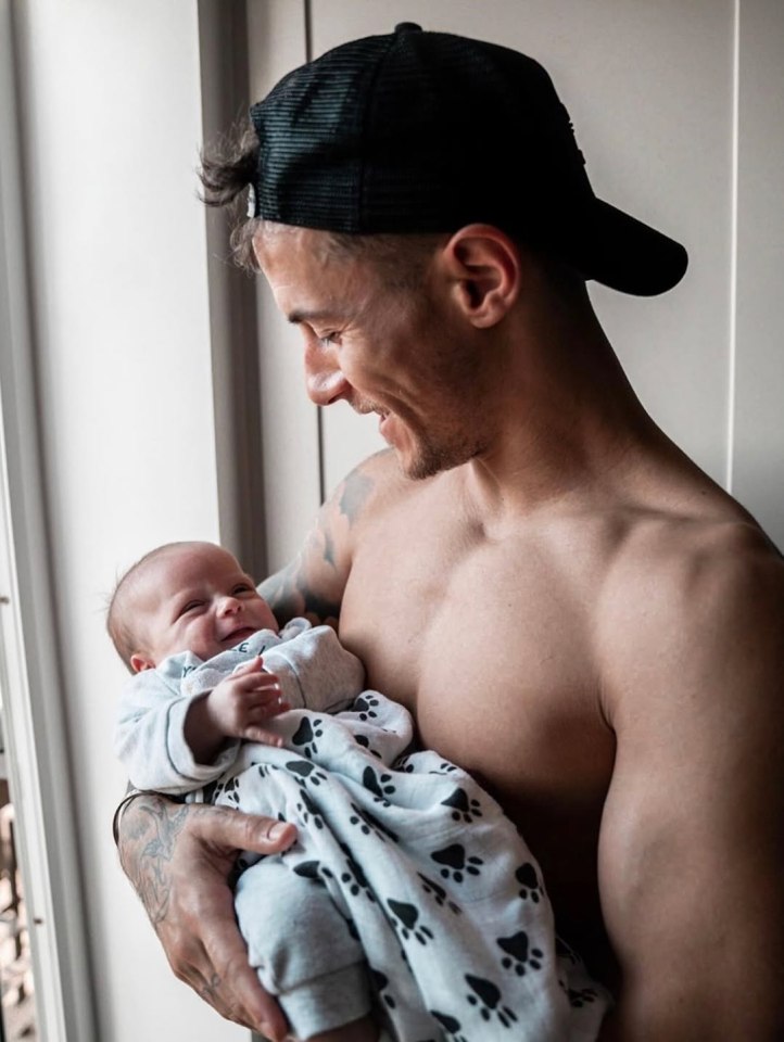 A shirtless man holds a baby wearing a paw print onesie.