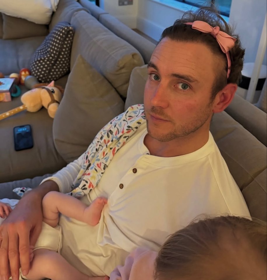 A man wearing a pink headband sits on a couch holding two babies.