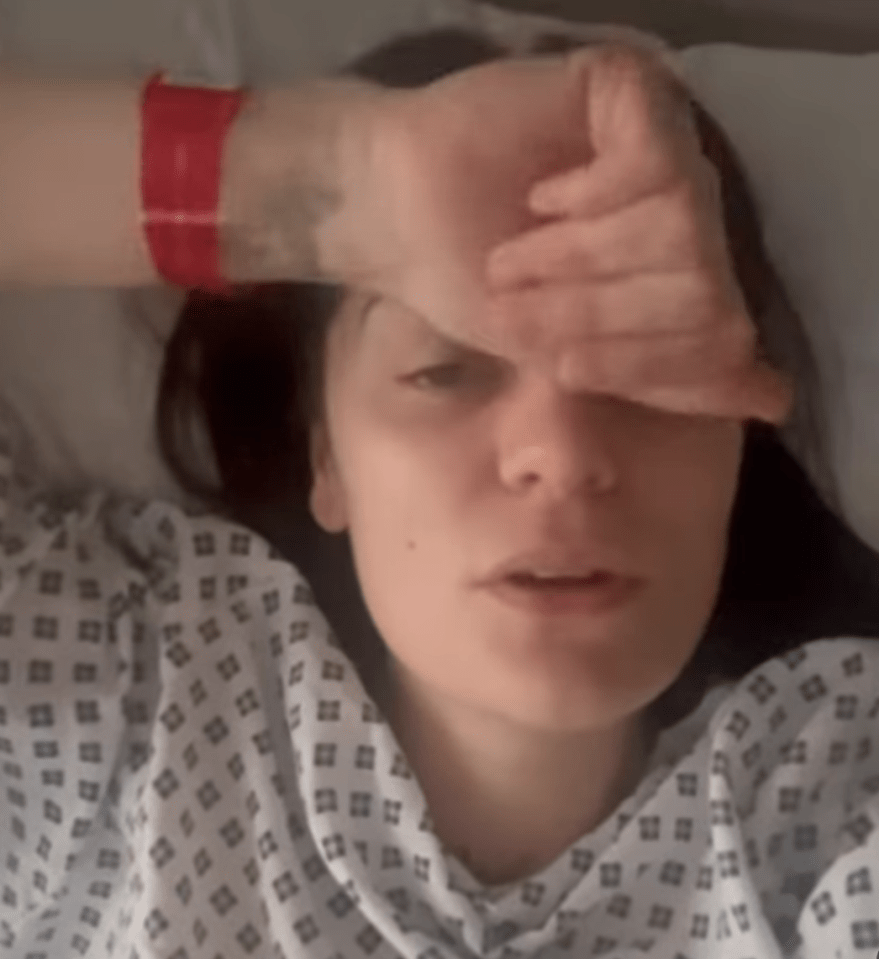 Woman in hospital gown lying in bed, hand on forehead.