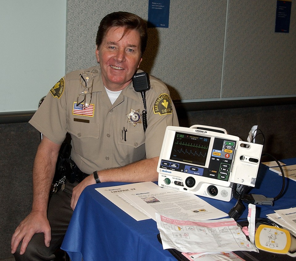 A sheriff sits at a table with a defibrillator and related documents.
