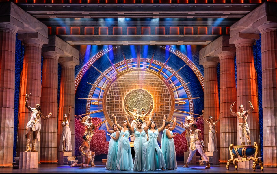 A stage production featuring a group of singers in light blue gowns and dancers in gold and white costumes, set against a backdrop with large columns and a circular design.