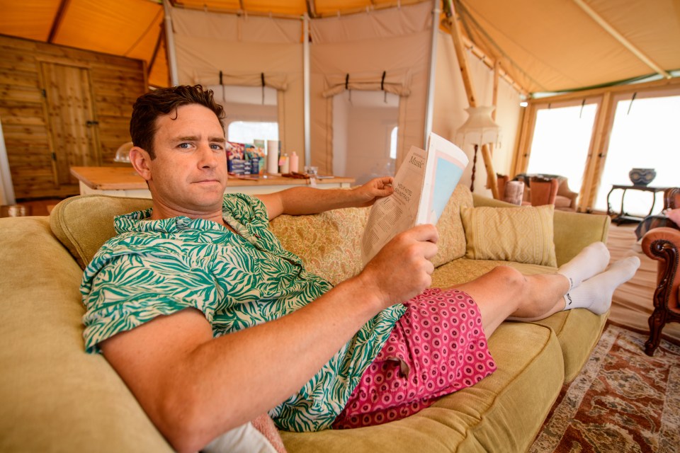 Man relaxing on a couch inside a tent, reading a newspaper.
