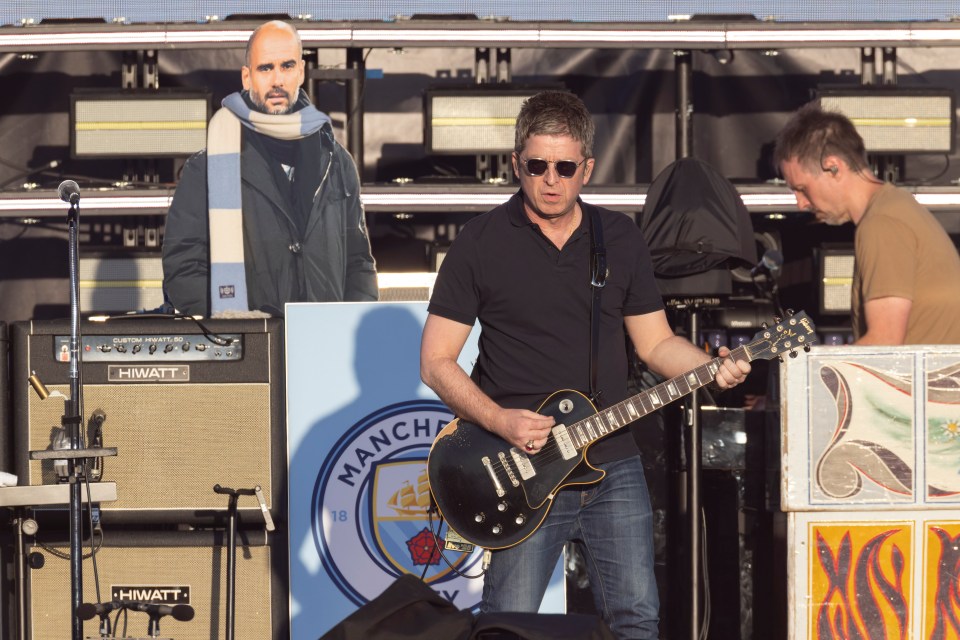 Noel Gallagher playing guitar at a concert with a Pep Guardiola cutout in the background.