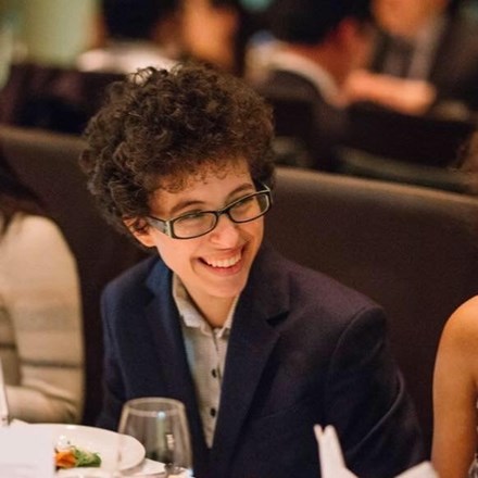A young person with curly hair and glasses smiles at a table.